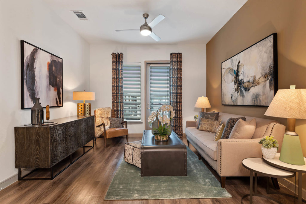 Living room with door to the patio/balcony, wood-like floors, ceiling fan, bronze accent wall and modern furniture