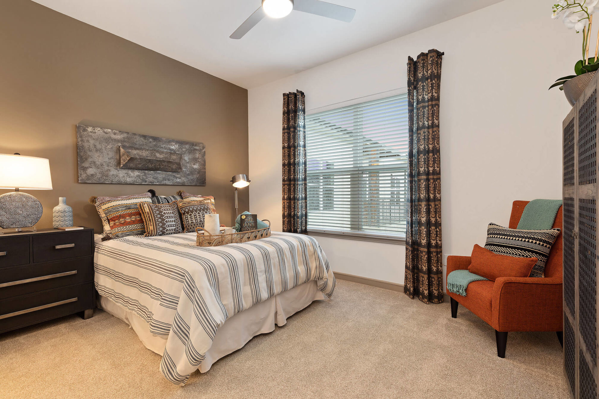 Bedroom with tan accent wall, large window, queen sized bed, large dresser, plush carpet and ceiling fan